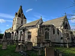 Side view of light coloured stone church with two stepped ranges ending in tower to left against a medium blue sky with white clouds on a sunny day