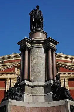 Photograph of an elaborate memorial centered on a massive stone column. At the base of the column there are two bronze statues of seated figures. There is a bronze statue of a man standing on top of the column. There are extensive carved inscriptions on the column itself. Behind the memorial is the brick and stonework facade of a large and elaborate building; there is a clear blue sky above the building.