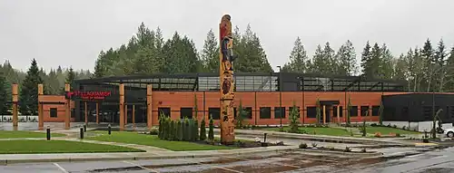 A large cedar building with a glass ceiling. There is a large totem pole near the entrance, with a parking lot in the foreground