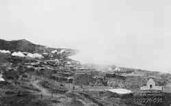 A beach crowded with military stores. Near the foreshore a plume of smoke rises