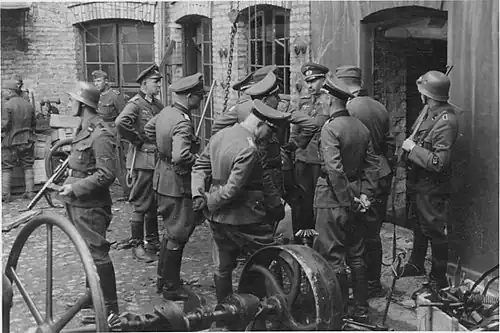 NARA copy #3, IPN copy #3 Discussing the evacuation of the factory The guard on the left is Josef Blösche. The photo depicts the Herman Brauer helm repair shop at Nalewki 28–38. 24 April 1943