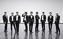 Black and white picture of nine men (members of Super Junior) wearing tuxedos are standing in a line