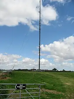 A large antenna stretches into the sky. It sits on a green field surrounded by buildings.