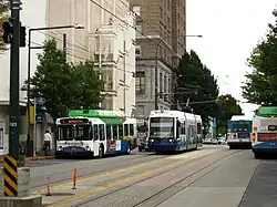 A streetcar and several short buses seen sharing a four-lane street with overhead wires and streetlamps.