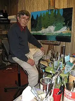 Ted Smith, an elderly white man in glasses, sits on an office chair in front of an easel with a landscape of rocks and trees on it. He is surrounded by paint brushes.