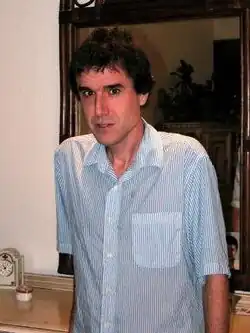 A middle-aged man, with short-black hair, in a dress shirt, looks at the camera. In the background, a dresser and a mirror are displayed.