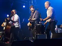 A side angle photo of a band playing in front of an audience under blue tinted lights. Two band members are playing string instruments, one is playing an accordion, and one is playing the drums.