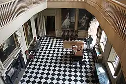 A photograph looking down on the interior of the Great Hall, showing the chequerboard floor tiles, several of the large wall portraits and pieces of furniture placed at the edges of the room.