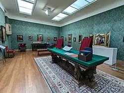 A large room with skylights and wooden floors, displaying a range of paintings on the walls and on tables
