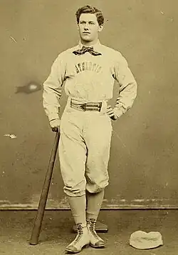 A baseball player is standing, facing the camera, with the top of a baseball bat in his right hand, and the bottom resting on the ground