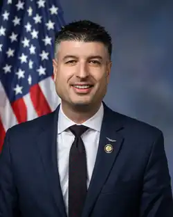 Barrett smiling in front of the U.S. flag, wearing a dark blue suit with an Army Aviator Badge, his House member pin, a white shirt, and a black tie with red dots.