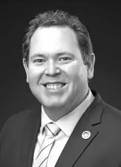 A monochrome portrait photo of a man with short hair, 5 o'clock shadow, wearing a suit coat and tie; he is looking and facing the center of the camera with a happy expression.