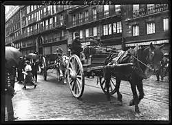 Line of horse-drawn carts moving up a street.