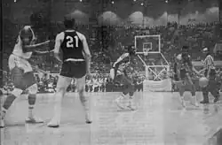 Two men are on a basketball court, one with his back to the camera and one dribbling a ball toward the camera in a black and white photograph.