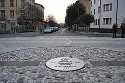 Metal plaque installed in 2004 in the middle of the square at the border point, once crossed by the "Gorizia Wall&nbsp;[it]" [the latter separated Slovenian Yugoslavia from Italy, and during this era border controls were carried out the same way as along Berlin wall, moreover this was strengthened with barbed wire to prevent escapes [citation needed]]