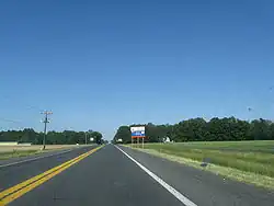 A two-lane road passing through farm fields. A blue and white sign on the right indicates an ongoing construction project on US&nbsp;113.