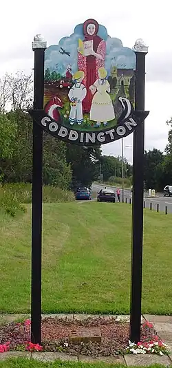 The word Coddington beneath a painted sign featuring a woman with two children in old fashioned dress against a contrived rural backdrop with a church, a working tractor, a world war two bomber, a fox and a badger standing tall atop two slender posts