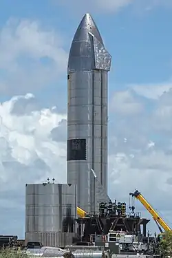 Photograph of a large, mostly featureless silver rocket at a facility