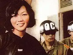 A Vietnamese woman dressed in black smiling on the left, next to two guards on the right wearing military uniforms and helmets.