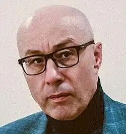 Colour photo portrait of an unsmiling, bald, bespectacled man with strong features, and designer stubble.