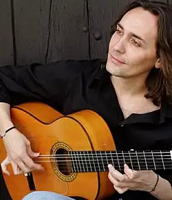 A man wearing a black shirt is holding a Spanish guitar.