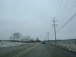 Westbound IL 120 just west of city of McHenry and narrowing to two lanes. Riding on concrete, there are open areas on both sides of the road with scattered trees on the left and utility poles on the right.