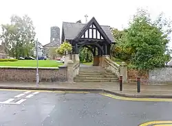 Dark timber half structure with tiled roof and white vertical infill panels close to road having lawns and path towards the church in background against a light sky