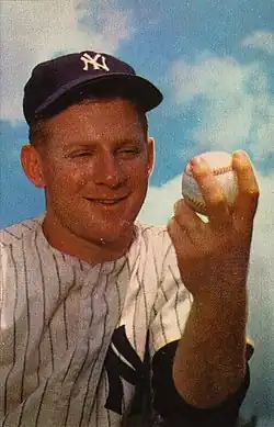 A smiling man in a pinstripe baseball jersey and dark baseball cap with an interlocked white "NY" on the front, holding a baseball