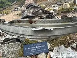 A small yellow plush mascot figure is placed inside a cracked piece of metal wreckage. The figure has a smiling face and is dressed in a dark outfit. The ground around it is rocky, with small plants and other pieces of metal debris scattered nearby. A red ribbon or flower is attached to the wreckage in front of the figure.