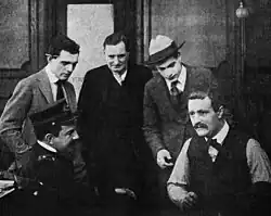 Black and white film still showing a man with a mustache being questioned