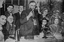 Black and white photo. William Hale Thompson stands before a desk, surrounded by others, while using a telephone of the candlestick telephone-design, with the mouthpiece held to the right-side of his face by his right hand and the earpiece held to his left ear by his left hand