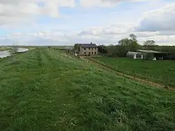 Colour photograph showing a small river on the left, and grass with a cottage on the right