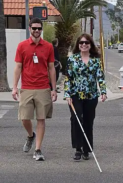 A man and a woman crossing the street. The woman is using a white cane