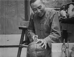 A black-and-white image of a grinning man cutting a melon with a saw