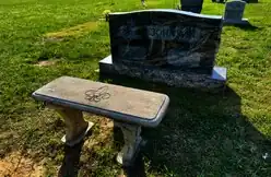 A glossy, dark-colored, and marbled headstone sits in a grassy cemetery, accompanied by a concrete bench engraved with a fleur-de-lis